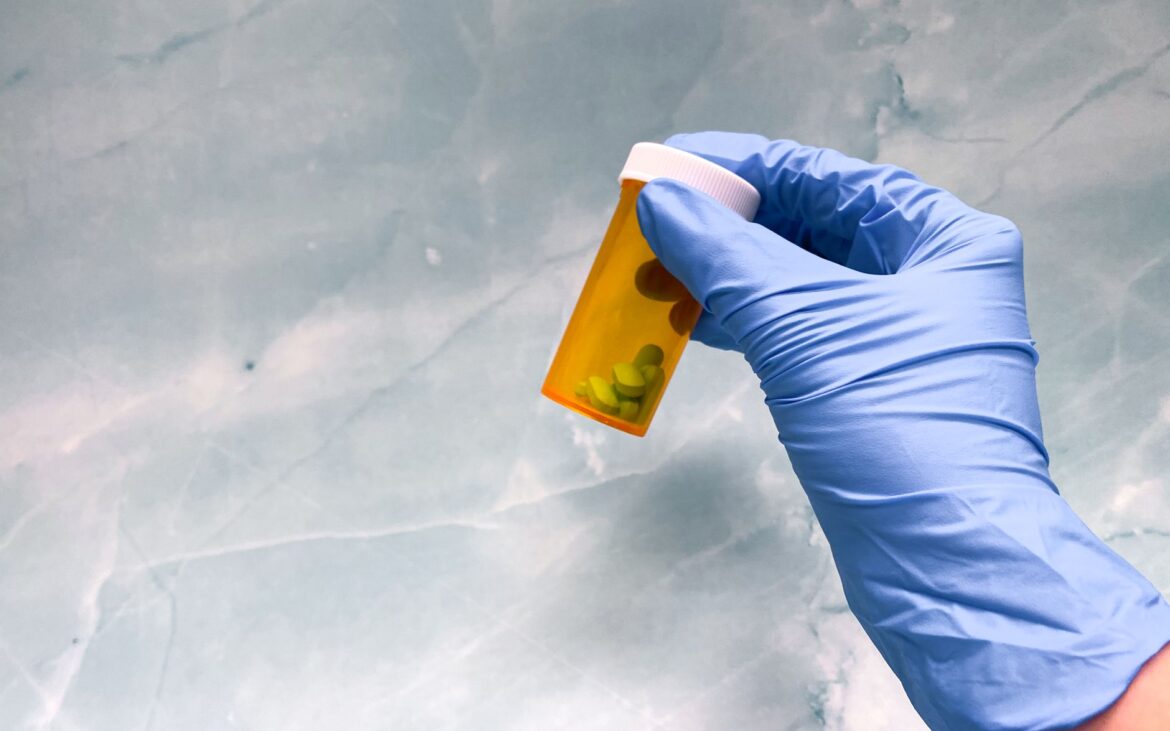 A gloved hand holds a prescription pill bottle against a textured background.