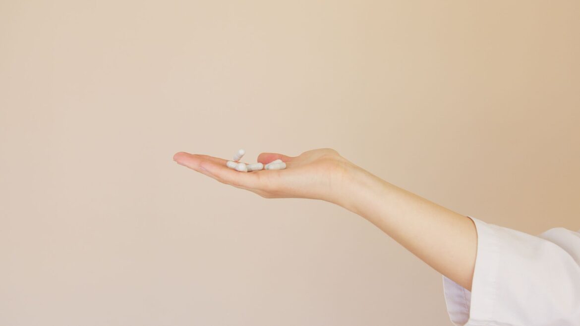 Side view of crop unrecognizable female pharmaceutist in white uniform with  pile of white painkillers on palm against light brown background