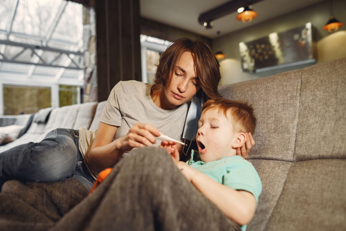 Calm woman in jeans watching digital thermometer while sitting near little yawning boy covered with blanket on cozy couch in afternoon at home